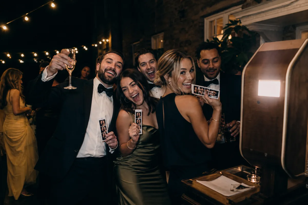 Illustrative evening wedding reception scene with guests posing and holding photo strips.